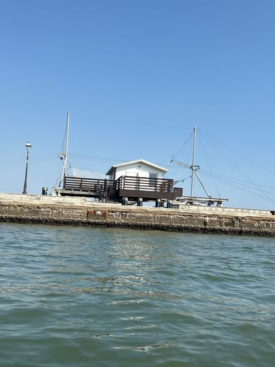 This image shows a fishing house on stilts in Cesenatico, Italy, with the Adriatic Sea in the foreground. The fishing house is a white, two-story structure with a wooden deck and railings. It is supported by dark blue stilts. A large, mast-like structure with a net and cables rises from the house, used for fishing. There is a lamppost to the left of the house. A bicycle is parked on the deck. The house sits on a stone embankment that lines the water. The water is choppy and a clear blue sky is visible above. The overall scene is one of a functional, traditional fishing setup in a coastal town.