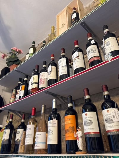The image depicts an interior view of a wine shop in San Gimignano, Italy, showcasing three stacked shelves filled with bottles. The background reveals a light-colored, rough-hewn stone wall and a plain ceiling.

The topmost shelf features several dark-glass wine bottles, with some positioned inside open wooden crates branded "MARRELLI." A single ornate gold-colored bottle stands prominently. To the far left, a potted plant with pink geranium flowers is visible.

The middle shelf contains numerous dark and light-glass wine bottles. Many bottles are adorned with white paper price tags, some legible with details such as "Chianti Classico" and "Carmignano." A small, terracotta-colored dish holding a dark, dried floral object is centrally located on this shelf.

The bottom shelf displays a collection of wine bottles, primarily dark-glass, with labels identifying Italian wines like "Morellino di Scansano" and "Capanneto." Several bottles carry price tags, one clearly indicating €19.00. A small figurine of a chef or baker, dressed in a white uniform and a black mask with hands clasped, stands upright on this shelf. A cluster of wine corks is visible next to a bottle on the lower right. No individuals are present in the scene.