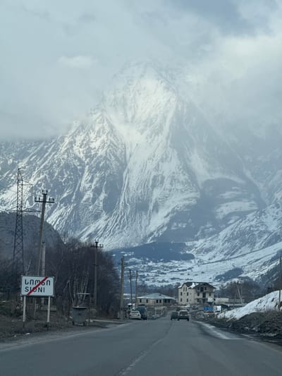 The image displays a mountainous winter landscape in Sioni, Georgia, observed from a paved two-lane road. In the immediate foreground on the left, a white rectangular road sign with "ს ი ო ნ ი" and "SIONI" written in black, crossed by a diagonal red line, signifies departure from the locality. Next to the sign stands a grey, wheeled trash bin. Multiple utility poles with power lines are distributed along both sides of the road, and a tall metal lattice power tower is visible on the far left.

In the midground, several buildings, including residential structures and some seemingly under construction, are clustered within a snow-dusted valley. Four vehicles are present on the road: three passenger cars are heading away from the viewer, while one darker vehicle is parked on the left shoulder. Bare trees and sparse vegetation are scattered throughout the scene.

The background is dominated by towering, rugged, snow-covered mountains, partially obscured by dense grey clouds that blend into the overcast sky. The environment suggests cold, wintry conditions.