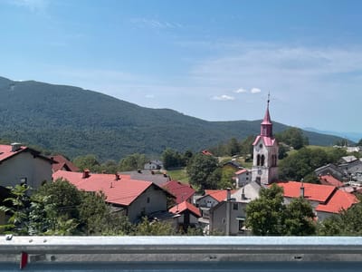 The image shows a view of the village of Col, Slovenia, from a higher vantage point, likely a roadside overlook. In the foreground, a silver guardrail with red reflectors is visible across the bottom of the frame. Beyond the guardrail, the village of Col is nestled in a valley. The houses are characterized by their red-tiled roofs and are surrounded by lush green trees. Prominently featured in the village is a white church tower with a pink spire, topped with a cross. The village is situated at the base of a large, densely forested mountain that dominates the left side of the image. The mountain slopes upwards towards a clear blue sky with a few scattered white clouds. The overall scene depicts a tranquil rural landscape on a sunny day.