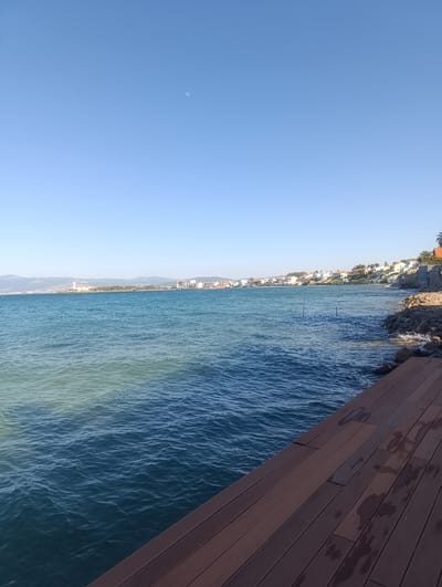 The image depicts a serene coastal scene in Urla, Turkey. The clear blue sky dominates the upper portion of the frame, with a faint crescent moon visible. Below, the tranquil blue sea stretches towards the horizon, meeting a distant shoreline lined with buildings and hills. The water exhibits varying shades of blue and green, suggesting shallow areas or differing depths.

In the foreground, a wooden deck runs diagonally across the bottom right of the image. The planks are a rich brown, some showing evidence of dampness, hinting at recent rain or proximity to the sea. The deck provides a vantage point overlooking the water.

Along the shoreline, a row of houses with white walls and terracotta roofs creates a picturesque Mediterranean village. The coastline is rocky in places, with waves gently lapping against the shore. The overall impression is one of peacefulness and natural beauty.

The time of day appears to be late morning or early afternoon, given the bright sunlight and clear sky. The weather is fair and calm, with no strong winds evident in the water. There are no people or specific activities taking place, giving the scene a quiet, contemplative feel. The location is identified as Urla, Turkey, which is a coastal town known for its scenic beauty. No visible text is present in the image.