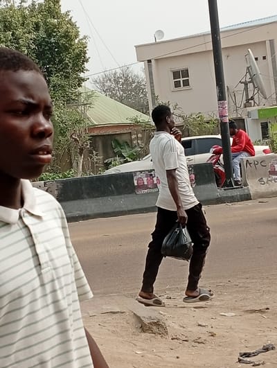 The image depicts an outdoor urban scene in Darmanawa, Nigeria. In the foreground, a young male with dark skin and short dark hair is partially visible, facing left with a neutral expression. He wears a light-colored polo shirt with thin horizontal green stripes.
In the mid-ground, a male with dark skin, wearing a white short-sleeved t-shirt, dark trousers, and light-colored sandals, walks from left to right across uneven dirt ground adjacent to an asphalt road. He carries a black plastic bag in his left hand, and his right hand is raised to his chin area.
Further in the background, a third male with dark skin, dressed in a red long-sleeved shirt and blue jeans, is seated on a low concrete road barrier. A red scooter or motorcycle is parked near the barrier.
The environment includes a dark utility pole, positioned between the walking male and the seated male, with several small posters affixed, one bearing "APC" text. Behind the barrier, a white sedan is parked. Multiple buildings are visible: a multi-story light-colored building with a flat roof and a satellite dish, a light-colored building with a single square window, and a building with a green sloped roof. Various trees and bushes are present, particularly behind the structures. The ground in the immediate foreground consists of rough, sandy dirt.