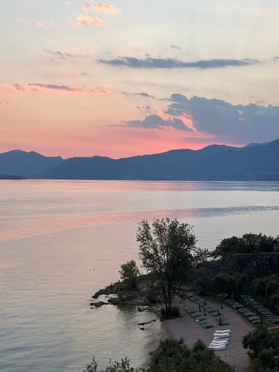 This is a photograph of a sunset over Lake Garda, viewed from the town of Garda, Italy. The sky is filled with soft pink and orange hues, with scattered clouds. In the distance, the silhouette of rolling hills lines the horizon. The lake itself is calm, reflecting the colors of the sky, with gentle ripples on its surface. A small, rocky shore juts out into the water on the right side of the frame. Beyond the shore, a sandy beach is laid out with rows of empty lounge chairs and umbrellas, suggesting a resort or public area. Several white inflatable rafts are stacked neatly on the sand. Trees and bushes dot the landscape, with a prominent tree near the center of the image. The overall scene depicts a tranquil and picturesque evening on the Italian lake.