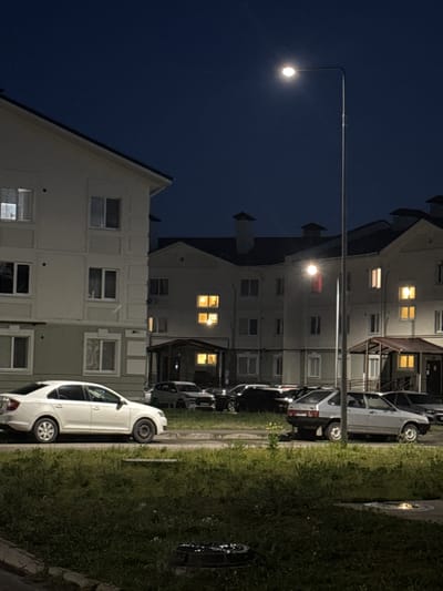 This is a photograph taken at night in the city of Kuyuki, Russia. The scene depicts a residential area with multi-story apartment buildings and parked cars. A street light illuminates a portion of the image. The sky is dark blue, indicating it is nighttime. The buildings have windows, some of which are lit from within, suggesting occupancy. Several cars are parked in front of the buildings, on what appears to be a paved area bordering a grassy strip. The overall impression is one of a quiet, residential neighborhood after dark.