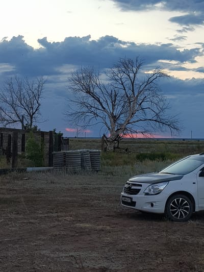 The image shows a white Chevrolet Cobalt parked on a dirt road in a rural area. The car is facing right and is partially visible in the foreground. Behind the car, there is a landscape with dry grass, a few bare trees, and some wooden structures. In the distance, there is a flat horizon with a cloudy sky. The sky has a mix of dark blue and light gray clouds, with a hint of pinkish-orange light visible on the horizon, suggesting either sunrise or sunset. There are no people visible in the image. The location is identified as the village district of Dolen, Kazakhstan.