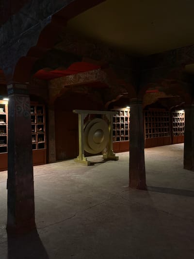 The image depicts an interior view of a dimly lit hall or covered corridor within the Cinema Museum (Musée du Cinema) in Ouarzazate, Morocco. The space features robust architectural elements, including multiple dark, weathered pillars supporting intricate arched structures. These arches and overhead beams are decorated with faded traditional patterns in red, dark blue, and earthy tones, while the ceiling itself is a muted yellow-orange. The floor is concrete, displaying visible cracks and signs of wear.
Centrally positioned in the midground is a large, golden-colored gong, composed of two concentric circular discs, mounted within a sturdy, matching golden frame. Along the rear and side walls, numerous cubbyhole-like display shelves are visible, each compartment containing small, dark, indistinct objects, possibly artifacts or film props. Some of these shelves are illuminated by internal lights, casting a soft glow on their contents and the immediate area. No individuals, actions, or interactions are present in the scene.