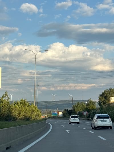 The image depicts a highway scene in the city of Berbuki, Georgia. Two white cars are driving on the road. In the background, there are several wind turbines on a hill, along with trees and utility poles. The sky is partly cloudy, with a mix of blue and white. The overall impression is that of a rural or semi-rural setting within or near the city.