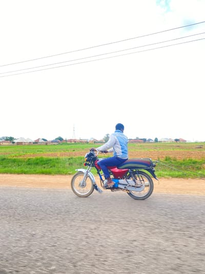 A young man is riding a motorcycle on a paved road in a rural area. He is wearing a blue and white jacket and blue pants. The motorcycle is red and silver. The road is lined with green grass and some buildings in the background. The sky is bright and clear, suggesting it is daytime. The image appears to be taken in a village in Nigeria, specifically Ladura Foron. There are power lines visible in the distance. The man appears to be enjoying himself while riding his motorcycle.