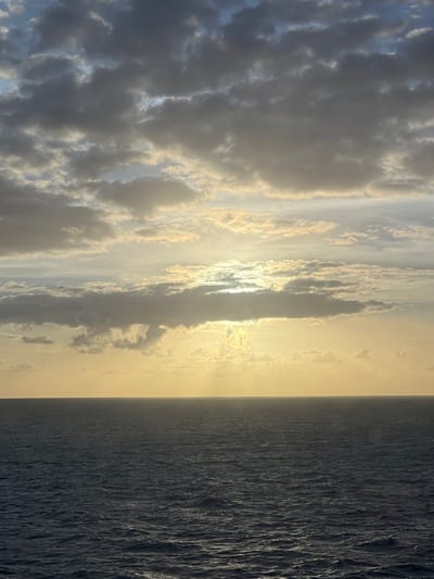 An overcast sky with the sun peeking through the clouds illuminates a vast expanse of dark, choppy ocean. The sun's rays create a glowing effect on the horizon. The sky is a mix of grey and white clouds with hints of blue. The ocean's surface is textured with small waves, reflecting the muted light from above. The scene is devoid of any human presence or man-made structures, suggesting a remote or open-water location. The overall impression is one of a serene, natural landscape, possibly at dawn or dusk.