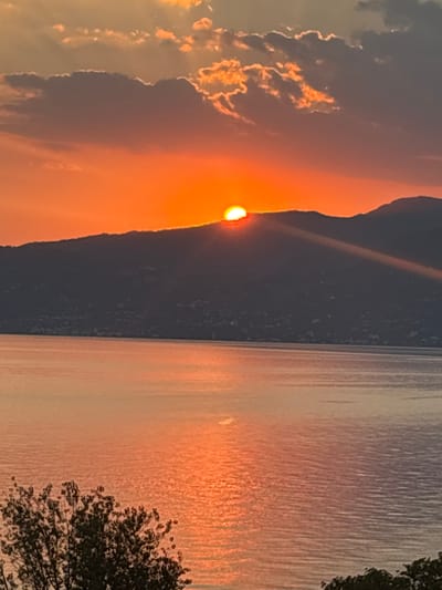 The image depicts a sunset over Lake Garda, Italy. The sun is partially obscured by a dark, silhouetted mountain range, casting vibrant orange and yellow rays across the sky. The calm water of the lake reflects the warm hues of the sunset. In the foreground, the dark foliage of a tree is visible on the right side of the frame. The scene is serene and devoid of any human presence.