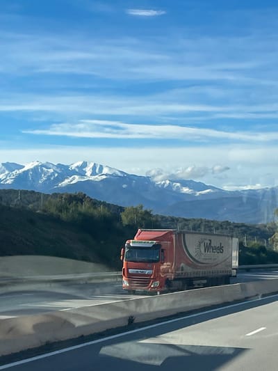 A vibrant blue sky with elongated clouds contrasts sharply with snow-capped mountains. An orange truck with "Wheels" printed on its trailer travels down a highway. The roadway is reflective, showing a hint of scenery and vehicle. Green trees line the edge of the road, positioned between the mountains and the highway.