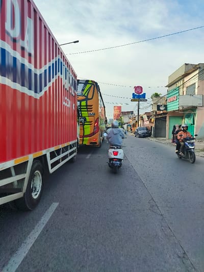 The scene shows a narrow street in Bojongsoang, Indonesia, with a large red container truck parked on the left side of the road. The truck is partially blocking a yellow and green bus that is driving in the opposite direction. Behind the bus, a white and grey scooter is traveling on the right side of the road towards the viewer. The scooter has a person wearing a white helmet riding it. There are three pedestrians walking along the sidewalk on the right side of the road. The road is asphalt and there are buildings along the sides. There are power lines running above the street. There is a sign for "ME Bread" on the right side of the road. The sky is clear and blue. The time of day is daytime. The location of the scene is -6.971 latitude and 107.637 longitude. The scene is a typical street in Bojongsoang, Indonesia.