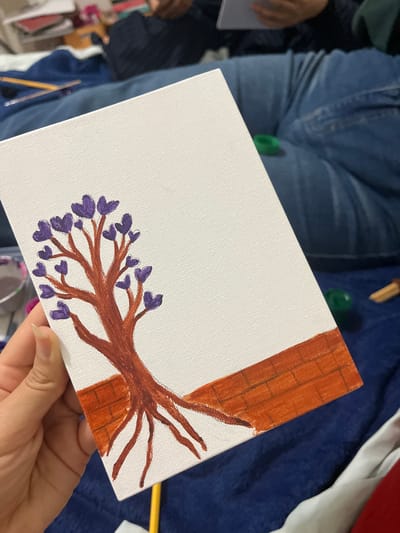 The image shows a close-up of a canvas painting of a tree with purple heart-shaped leaves and exposed roots, standing on a brick-like foundation. The painting is held by a person's hand, with fingers visible on the left side. The background is out of focus and appears to be a soft, dark blue fabric, possibly a blanket or clothing, with some other objects and people also out of focus. There is no discernible information about the location being in the city of San Luis, Peru.