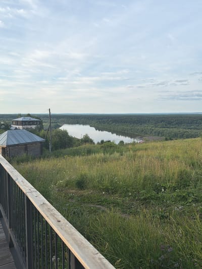 The image depicts a scenic overlook in Cherdyn, Russia, featuring a wide, calm river flowing through a dense, green forest. The foreground is dominated by a wooden railing and walkway, with tall, wild grasses and scattered wildflowers on a hillside. Two rustic wooden structures, resembling traditional Russian houses or outbuildings, are visible on the left side of the frame, with one appearing older and more traditional than the other. A utility pole with wires stands between the structures and the river. The sky is partly cloudy with a soft, diffused light, suggesting it is either early morning or late afternoon. The overall impression is one of rural tranquility and natural beauty.