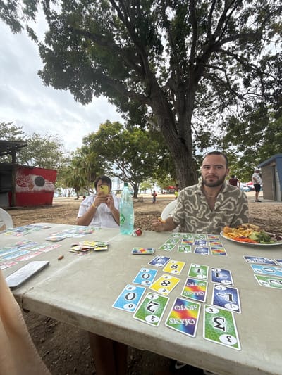 Here is a detailed description of the image:

**General Scene:** The image shows a group of people playing the card game SKYJO outdoors. The setting appears to be a park or recreational area with trees and some structures in the background.

**People:** There are at least two people clearly visible in the image. One person is looking directly at the camera with a slight smile. They are wearing a patterned shirt and jewelry. Another person is sitting with their face towards a smartphone. The rest of the image is too blurry to gather other information about the people.

**Objects:**
*   **Cards:** A large number of SKYJO cards are laid out on a table.
*   **Food:** A plate of food (possibly a salad and fried items) is visible on the table.
*   **Drinks:** A bottle of water is present.
*   **Table:** The game is being played on a stone table.
*   **Structure:** A red and white storage shed (could be for food and drinks) with words "Silence" can be seen in the background.

**Environment:**
*   There are trees visible throughout the scene.
*   The ground appears to be sandy, suggesting the location may be near a beach.
*   The sky is partly cloudy.

**Location:**
There is not enough information to discern the location, the city of Port-Louis, France.