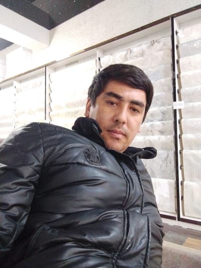 A male subject is depicted indoors in a commercial setting in Balykchi, Uzbekistan. The subject, an adult male with short dark hair, dark eyebrows, and a light to medium complexion, is seen from the chest up, looking directly at the camera with a neutral expression. He is wearing a black, shiny, puffer-style jacket with a high collar and a dark, circular emblem on the left chest. The background consists of vertical display shelves containing multiple white, plastic-wrapped items, possibly rolls of fabric or mattresses, stacked horizontally in compartments. A small label displaying '037' is affixed to a shelf divider. Above the displays, a white wall and a dark ceiling section with light-colored speckles are visible. The flooring appears to be wood or laminate.