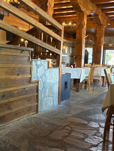 This media file captures the rustic and traditional interior of a multi-level building, likely a restaurant, hotel, or guesthouse, in Leshten, Bulgaria. The scene is devoid of people, suggesting it is unoccupied or awaiting guests.
The dominant features are the extensive use of natural materials, particularly heavy, rough-hewn wooden beams, wooden paneling, and irregular flagstone flooring and walls. Large wooden beams serve as structural elements and decorative features, with some exhibiting carved X-shaped patterns. Antlers or horn-like decorations are placed on several of these beams, enhancing the rustic aesthetic.
A prominent wooden staircase with open risers and sturdy railings leads to upper levels, hinting at the building's multi-story design. Tables, set with white tablecloths and some with checkered overlays, are visible, accompanied by distinctive wooden chairs, some featuring a slatted, bentwood-style design. Bottles containing red liquid are arranged on one table. A dark, rectangular object, possibly a speaker or heating unit, rests on a stone base adjacent to a dining area. A decorative metal light fixture hangs from a wooden beam.
Natural light filters through windows visible in the background, suggesting it is daytime. The overall ambiance is warm and inviting, characterized by the natural tones of wood and stone. There is no visible text in the image.