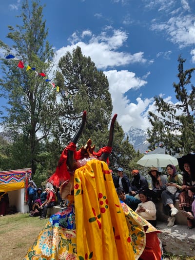 The image depicts an outdoor cultural event in Stingri, India, under a bright, partly cloudy sky, suggesting midday. In the foreground, a person in a traditional, elaborate costume, likely part of a performance, is central to the frame. The costume features vibrant yellow fabric with colorful circular patterns, a red headdress with prominent black horns, and intricate blue and gold detailing on the lower garment.
Behind the performer, a diverse group of onlookers is seated on a stone ledge. They appear engaged with the event, with some holding umbrellas for shade, including a large white one and a smaller black one. The onlookers are dressed casually, some wearing hats and headscarves. Their expressions are varied, but there's a general sense of observation and enjoyment.
The setting is a natural landscape with tall, mature trees framing the scene. In the background, a majestic snow-capped mountain range is visible, indicating a high-altitude location. Prayer flags in various colors are strung between trees, adding to the cultural ambiance. To the left, a small tent or canopy with colorful, patterned fabric is partially visible, suggesting a designated area for the event or performers.
The overall atmosphere is festive and communal, blending cultural performance with a scenic natural backdrop. The bright sunlight and scattered clouds indicate pleasant weather. There is no visible text in the image.