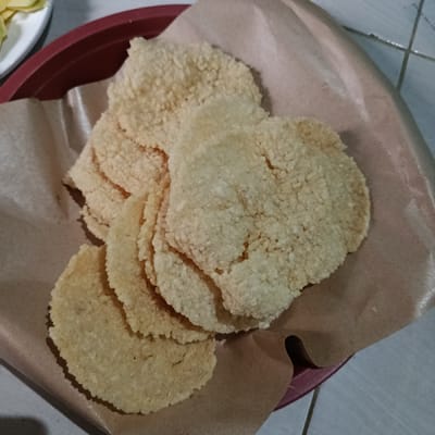 A plate of round, tan-colored crackers rests on brown parchment paper inside a maroon-colored dish. The crackers have a bubbly, textured surface and vary in shade. Some appear lighter and more puffed up, while others are flatter and darker. A partial view of a plate with food is visible in the upper left corner. The crackers fill most of the frame and appear to be the main focus.