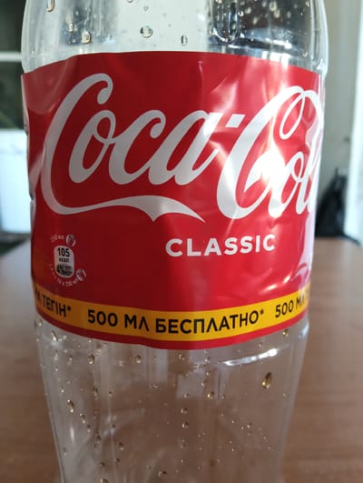 This is a close-up, eye-level photograph of a clear plastic bottle of Coca-Cola. The bottle is partially filled with a brown liquid and has condensation on the outside, indicating it is cold. The iconic red Coca-Cola label is prominent, featuring the white Coca-Cola logo. Below the logo, the word "CLASSIC" is printed in white.

On the left side of the label, there is nutritional information in Russian: "250 мл 105 ккал" (250 ml 105 kcal) and "47% РСС" (47% RDI), along with a breakdown of sugar content. A yellow band runs across the lower part of the label with text in Russian that reads "М.ТЕГІН* 500 МЛ БЕСПЛАТНО* 500 MA" (M. TEGIN* 500 ML FREE* 500 MA).

The bottle is placed on a wooden surface, likely a table, which is slightly out of focus. The background is blurred but suggests an indoor setting with natural light, possibly near a window, as indicated by the bright highlights and reflections on the bottle's surface. The overall lighting is bright, casting a slight shadow from the bottle onto the surface. There are no people visible in the frame. The image appears to be taken during the daytime.