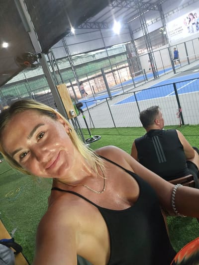The image shows a selfie taken by a woman with blonde hair and light skin. She is wearing a black tank top and a gold necklace, and her skin appears sweaty. Her expression is a slight smile. In the background, a man in a black sleeveless shirt is visible from the back, seated on a wooden bench. The setting appears to be an indoor sports facility with a green artificial turf floor, a blue and gray court, and metal support structures for the roof. A large fan is mounted on a pole in the background. A billboard with text and images is displayed on the far wall. The location is identified as Binh Thuan Ward, Vietnam.