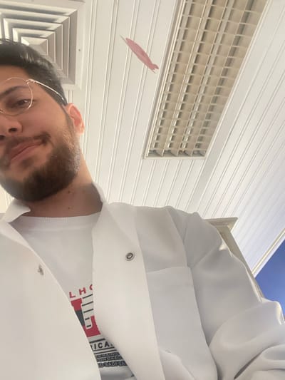 A male individual is depicted in an indoor setting in قرية الضبية, Syria, captured from a low-angle perspective. The subject's face is partially visible in the upper left, featuring dark hair, a short beard, and round, light-colored framed glasses. He displays a slight smile. The individual is wearing a white lab coat with visible metal snap fasteners, worn over a white long-sleeved shirt with a partially obscured graphic design in red, white, and black on the chest. The ceiling, constructed of white, horizontally paneled material, features a rectangular fluorescent light fixture with a grid diffuser on the right and a square air vent on the left. A small, pink, stylized airplane-shaped object is suspended from the ceiling between the subject and the light fixture. Portions of a light-colored structure and a blue wall or background element are visible on the right side of the frame.