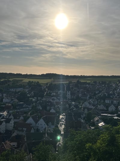 This high-angle outdoor photograph depicts the municipality of Untergruppenbach, Germany, under bright sunlight. The upper portion of the image features a radiant, overexposed sun positioned in the upper-center, generating a distinct lens flare. The sky is partially covered by scattered, thin clouds and traversed by multiple linear contrails from aircraft, with one prominent contrail extending vertically in the upper right quadrant.

The midground showcases the dense residential area of Untergruppenbach, characterized by numerous houses predominantly featuring gabled roofs with reddish-brown, dark gray, and white tiles. A centrally located church with a tall, dark spire and a white clock tower is a prominent architectural feature. A winding road is visible traversing the town, with several parked and moving vehicles. A distinct green lens flare, possibly originating from a street light, is present in the lower-middle portion of the town, slightly to the left of the main road. Some rooftops display solar panels.

Beyond the town, the background reveals agricultural fields in various shades of green, indicating different cultivation patterns, bordered by dense forested areas that form the horizon. The immediate foreground is occupied by dark green tree foliage, partially obscuring the lower sections of the town. No identifiable individuals are visible within the frame.