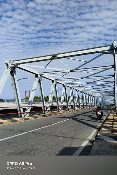 The image shows a steel truss bridge in Neuboknaleung, Indonesia. A line of motorcycles are on the bridge. The bridge has red railings and yellow-and-black striped borders. The sky is light blue with scattered white clouds. The horizon shows trees on either side of the river.