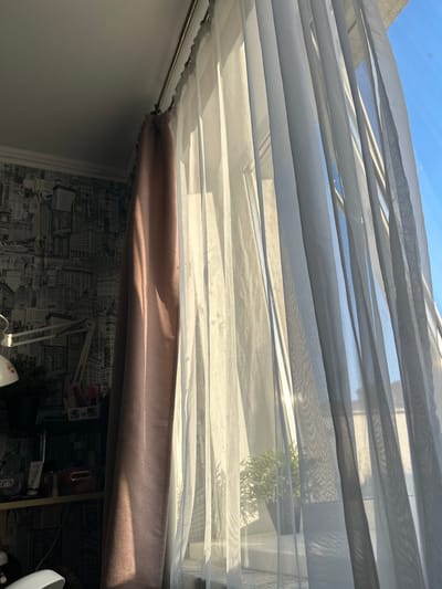 The image shows a view looking upwards through layered sheer white curtains towards a blue sky. A brown curtain valance is visible in the midground. The background includes a wall covered with a black and white cityscape wallpaper. Beneath the valance is a cluttered desk with various objects including a lamp, plants, and desk accessories. The location details (Imeni Maksima Gor'kogo, Russia) are not discernible from the visual information provided in the image.