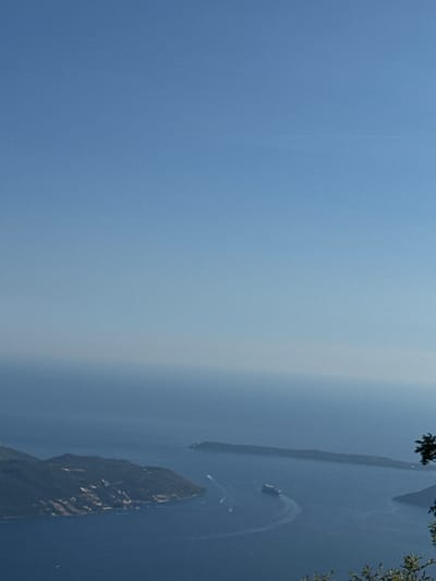 The image depicts a panoramic view of a coastal area, likely from an elevated vantage point. The upper portion of the frame is dominated by a clear, light blue sky. Below the sky, the sea stretches out, transitioning from a hazy, lighter blue near the horizon to a deep, rich blue in the foreground.

On the left side of the image, a lush, green, mountainous island or peninsula juts into the water. The slopes are covered in dense vegetation, and scattered along the coastline are small clusters of white buildings, suggesting a settlement. The topography of this landmass is rugged and uneven.

In the mid-ground, a long, narrow island or landmass stretches horizontally across the water. This island appears less vegetated than the one on the left, with some evidence of structures or development.

A large cruise ship is visible in the water, leaving a white wake as it navigates. Several smaller boats are also present in the water, some appearing to follow the larger vessel.

The environment is serene and expansive, characterized by the vastness of the sea and sky. The presence of the cruise ship and the coastal settlement suggests human activity within this natural landscape. The image captures a bright, clear day with good visibility. The specific location in Bunovići, Montenegro, suggests this is a view of the Adriatic Sea coastline.