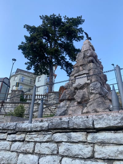 In Caprino Veronese, Italy, a monument stands against a clear blue sky, topped by a bronze eagle with outstretched wings. The monument is a rough-hewn stone structure, resembling a mountain peak, with a plaque inscribed "AI SUOI CADUTI 1959" (To its Fallen 1959). Two large metal projectiles, resembling artillery shells, are positioned on either side of the monument. A thick metal chain encircles the monument and its surroundings, attached to posts. Behind the monument, a large evergreen tree with dense foliage casts a shadow. To the left, a multi-story building with balconies and windows is visible, along with a lamppost. A stone wall, constructed from large, roughly cut stones, forms the foreground.