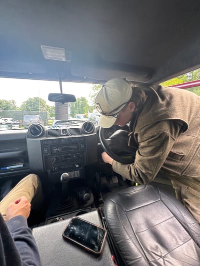 The image shows the interior of a vehicle, likely a Land Rover Defender, parked in Bramham, United Kingdom (latitude: 53.872, longitude: -1.38). A person wearing a brown tweed jacket, khaki pants, and a beige baseball cap is leaning over the driver's seat adjusting the steering wheel. Another person, only partially visible, is sitting in the passenger seat, wearing khaki pants and a navy blue jacket. A smartphone lies on the passenger seat. The vehicle's interior is black and features a black leather driver's seat, a black dashboard with various controls, and a gear shifter. The driver's side window is rolled down, revealing a view of the exterior environment, including green trees and other parked vehicles. The rearview mirror is also visible in the reflection.