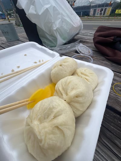 At a street-side picnic table in Dongmyeong-myeon, South Korea, a white styrofoam container holds a meal of steamed buns and yellow pickled radish. Two pairs of wooden chopsticks rest on the container, one pair piercing the pickled radish. Nearby, a white plastic bag, a brown jacket, and two small plastic dipping sauce containers are also visible. In the background, a road runs past several commercial buildings with signage in Korean.