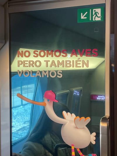 An upright, close-up shot captures a window, likely from a train or bus, featuring a whimsical scene with reflections and text. A cartoon-style pelican or stork figure, primarily white with orange wings, a long orange beak, and colorful striped legs, is attached to the right side of the window. It wears a small pink cap.
Prominently displayed on the window, reflected from the interior or applied as a decal, is Spanish text in a stylized, bold font. The text reads: "NO SOMOS AVES PERO TAMBIÉN VOLAMOS," which translates to "WE ARE NOT BIRDS BUT WE ALSO FLY," a clever play on words suggesting the vehicle's mode of transport.
To the upper left, a standard green emergency exit sign with a running person icon and a downward arrow is visible. Reflections on the window show distorted text, including "Wem A-10E" and "6110/T 30-A," originating from inside the vehicle. The view outside the window is obscured by glare or possibly a wintry scene with snow or ice, presenting a soft, light blue and white abstract background. The overall setting is consistent with a journey through Tres Cantos, Spain.