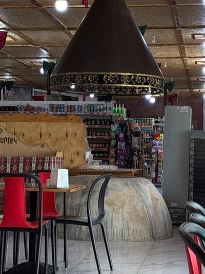 The image shows the interior of a market or small grocery store in Tsovagyugh, Armenia. The scene is dominated by various goods displayed on shelves, including canned foods, bottles, and packaged items. A large, conical ventilation hood hangs from the ceiling, decorated with ornate gold trim. In the foreground, there are tables and chairs, with two red chairs and two black chairs visible. A large, dome-shaped oven or display is situated in the middle of the room, with a wooden partition behind it. The overall ambiance suggests a casual, community-oriented establishment.