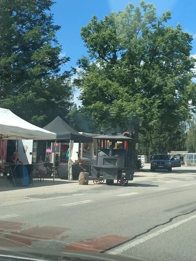 The image depicts an outdoor scene in Bližná, Czechia, on a sunny day with a clear blue sky. In the foreground, a dark grey, possibly metal, steam engine-like structure on wheels is parked on an asphalt road. It features a chimney emitting a light plume of smoke. To the left of the steam engine, there are two white and black tents, likely part of a market or event setup, with people visible inside and around them. Further to the right, a dark-colored SUV with its passenger door open is parked on the same asphalt area. In the background, dense green trees and some buildings can be seen. The overall setting suggests a public gathering or outdoor event.