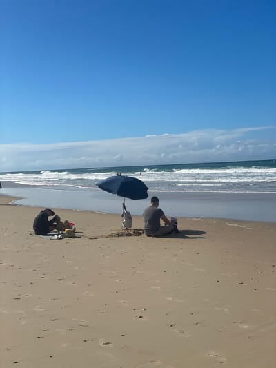 On a sandy beach in Surfers Paradise, Australia, two small groups of people are seated near the ocean under a clear blue sky. To the left, a person wearing a black jacket and cap is sitting on a grey mat with two small dogs. In the center, a dark blue umbrella shades a small mound of sand. To the right of the umbrella, a man wearing a grey t-shirt and light brown shorts is seated on the sand with a young child wearing a white hat. The ocean in the background has moderate waves, and there are a few distant figures in the water. The sand is marked with numerous footprints.