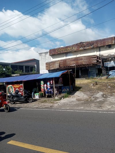 A daytime photograph depicts a street scene in Ban Phonhxavanh, Laos. The central feature is a dilapidated "Cipanas Theatre" building, identified by its rusted, ornate marquee with faded gold lettering, positioned in the background. The theatre's facade displays significant structural decay and rust. In the midground, a makeshift roadside stall with a blue tarp roof operates, positioned on a dirt and gravel patch in front of the abandoned theatre. The stall displays various packaged items, possibly snacks and drinks, and features a menu board on its left side. Two motorcycles are parked near the stall: a red scooter is partially visible in the left foreground, and a black scooter is parked directly in front of the stall. One individual, clad in light-colored attire, is partially visible from behind, interacting within the stall. The foreground consists of a paved road with a faded yellow line, indicating a traffic lane. Overhead, multiple power lines traverse a clear blue sky interspersed with white clouds. The area immediately surrounding the theatre building appears overgrown with dry vegetation and debris.