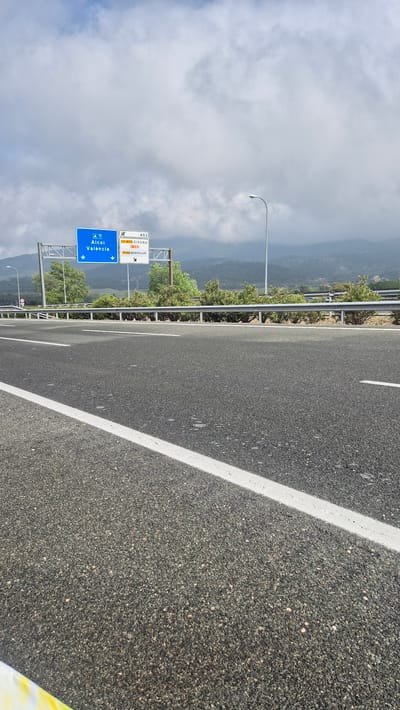 The image shows a stretch of the A-7 highway in Alcoi, Spain, leading towards Valencia. The road surface is asphalt with a painted white line marking the lane. A roadside sign indicates the direction to Alcoi and Valencia, along with other destinations like XIXONA and BENIFALLIM via different routes. To the left of the highway, a metal guardrail separates the road from vegetation and a view of the cloudy, mountainous landscape. A street light stands alongside the highway. The perspective suggests the photo was taken from the shoulder of the road or potentially from a vehicle moving along the highway.