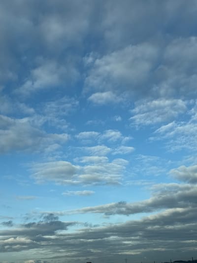 The image depicts the sky above Wolfisheim, France. The scene is dominated by cloud formations against a blue sky background. The upper two-thirds of the frame show a partially clear blue sky interspersed with scattered white to light grey cumulus and altocumulus clouds, some arranged in horizontal bands. The lower third of the image is characterized by a dense, continuous layer of darker grey stratocumulus clouds that extends to the bottom edge. No individuals, overt actions, or direct interactions are discernible. At the extreme bottom, faint, dark, indistinct silhouettes are visible, suggesting distant ground-level structures, including a possible utility pole-like element on the far right.