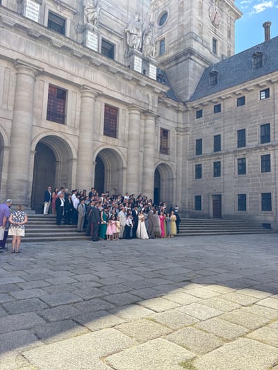 The image depicts a large group of approximately 60-70 individuals gathered on the stone steps leading to the entrance of the Royal Monastery of San Lorenzo de El Escorial, Spain. The central figures are a woman in a white wedding gown and a man in a dark suit, positioned towards the front of the group, suggesting a wedding party or formal celebration. Attendees are dressed in various formal attire including suits, dresses of differing colors such as pink, green, yellow, and grey. Most individuals are facing generally towards the camera, indicating a posed group photograph.

The background is dominated by the grand, multi-storied grey stone facade of the monastery, characterized by large arched doorways and windows with dark frames and some visible red-brown shutters. Prominent architectural features include large fluted columns, decorative sculptures, and inscribed plaques above the main arches. To the upper right, a clock tower with a visible clock face rises against a clear blue sky. A dark grey slate roof is visible on the right section of the building.

The foreground consists of a large, paved plaza or courtyard constructed from irregular, light grey stone blocks. Bright natural sunlight illuminates the scene, casting distinct shadows across the paved ground, originating from the left side of the frame. Two individuals, one in a purple shirt and the other in a floral top and white shorts, stand on the far left of the paved area, observing the main group.