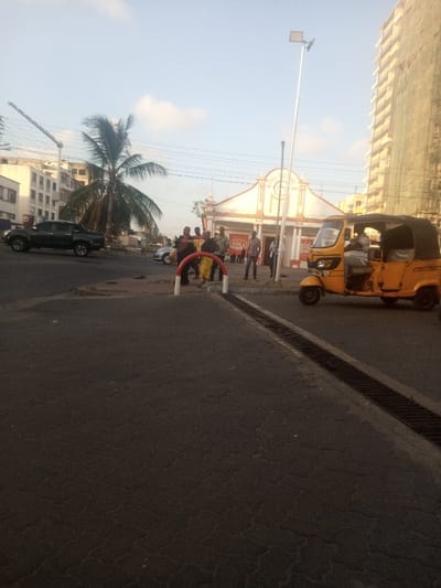 The image captures an urban street scene in Ganjoni/Shimanzi ward, Kenya, under bright daylight. The foreground features an asphalt road exhibiting minor surface cracking, with a metal drainage grate running along its right edge.
In the mid-ground, a group of approximately eight to ten individuals, dressed in casual clothing, are clustered near a red and white U-shaped barrier on a paved area adjacent to the road. One person within this group wears a yellow patterned garment. To the immediate right, a yellow three-wheeled auto-rickshaw, commonly known as a tuk-tuk, with a black canvas canopy, is positioned on the road. A male individual occupies the driver's seat. A tall, slender silver street light pole with an LED fixture extends vertically behind the tuk-tuk.
On the left side of the road, a dark-colored four-door pickup truck is visible, either stationary or in slow motion. Partially obscured behind the central group of people is a light-colored sedan. A prominent palm tree with green fronds stands between the left-side buildings and the central activity.
The background features several structures. Dominating the center-right is a distinctive building with a white and light brown/orange facade, characterized by an ornate arched entrance and a sign partially visible, reading "BU SAGA." To the far right, a tall, multi-story building under construction is enveloped in green scaffolding netting. On the far left, multiple multi-story buildings are visible, with a construction crane extending above one of them.