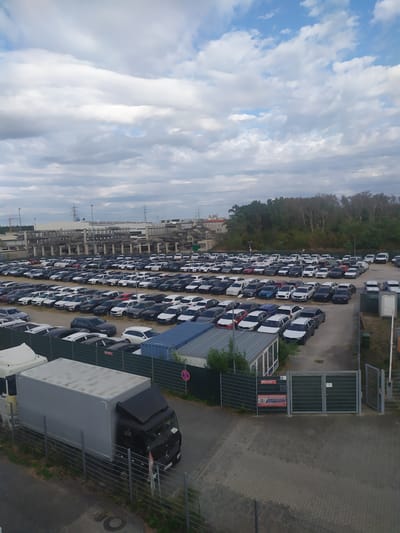 The image depicts an outdoor car storage lot, likely for a dealership or logistics company, in Bischofsheim, Germany. Rows upon rows of cars, predominantly sedans and SUVs, are neatly parked on a paved surface. The vehicles are varied in color, with a prevalence of dark and white models. A large, grey box truck with a black cab is parked in the foreground, partially obscuring a wire fence. The background features a mix of industrial structures, including what appear to be warehouses or parking garages, as well as overhead power lines and towers. A dense line of trees borders the lot in the distance, under a cloudy sky. A security gate with a "SECURITY" sign and a "KEINE FAHRT" (No Entry) sign is visible on the right side of the image. No people are visible in the frame.