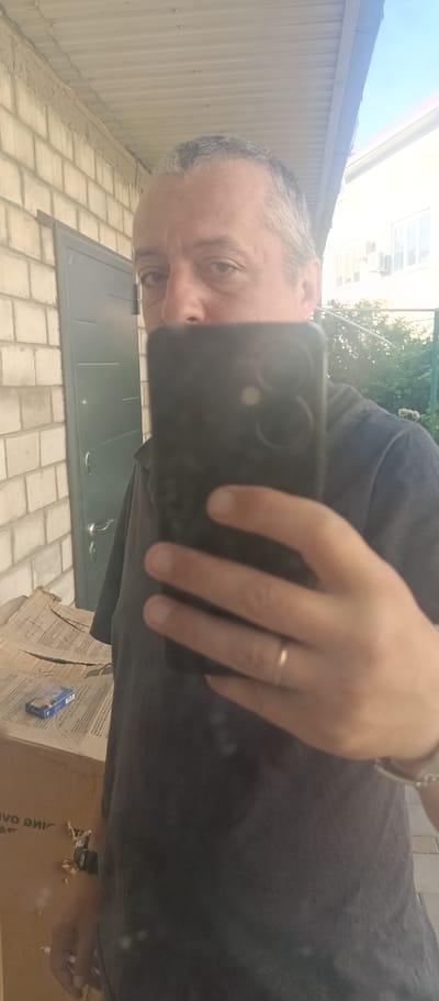 A man with short gray hair, wearing a dark t-shirt, takes a selfie in front of a brick wall and a green door. He is holding a black smartphone in his right hand, obscuring part of his face. A gold ring is visible on his left ring finger. To his left, a cardboard box is on the ground, with a pack of cigarettes on top of it. In the background, there is a building with windows and a green fence. The image appears to have been taken outdoors on a sunny day. The scene is set in Voskresenskiy, Russia.