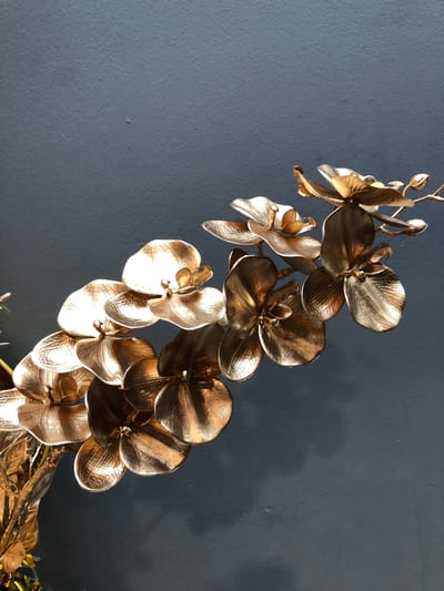 A close-up, eye-level, vertical shot captures a metallic gold artificial orchid branch against a dark grey, textured wall. The branch, adorned with numerous intricately detailed orchid blooms and buds, extends from the lower left corner towards the upper right. The orchid petals are rendered with a textured surface, mimicking the natural veining of real flowers, and they reflect light with a polished sheen. The background wall is uniformly dark and shows subtle imperfections in its texture. The lighting appears to be ambient, creating soft shadows and highlighting the metallic luster of the orchids. No people or other objects are visible. The scene is devoid of any context that would indicate a specific location within the city of Vaughan, Canada, or any other place.