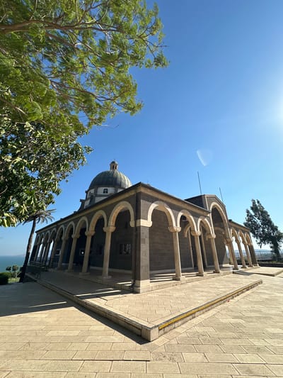 This media file captures a low-angle view of a domed building with arched colonnades, bathed in bright sunlight under a clear blue sky. Green trees frame the left side of the image, their leaves rustling gently. The building's exterior is constructed with dark stone blocks, contrasted by lighter-colored stone arches and pillars. A small dome, topped with a cross, is visible at the apex of the building. The ground in the foreground is paved with rectangular stone tiles, creating a textured surface that casts long shadows, indicating it is likely midday. The scene appears serene and peaceful, with no people visible. The location cues, such as the architecture and the presence of palm trees in the distance (partially visible), combined with the provided location context of Emek HaYarden Regional Council, Israel, suggest this is a significant religious or historical site in that region, possibly the Mount of Beatitudes. The bright, sunny weather and clear sky create a vibrant and uplifting atmosphere. There is no visible text in the image.