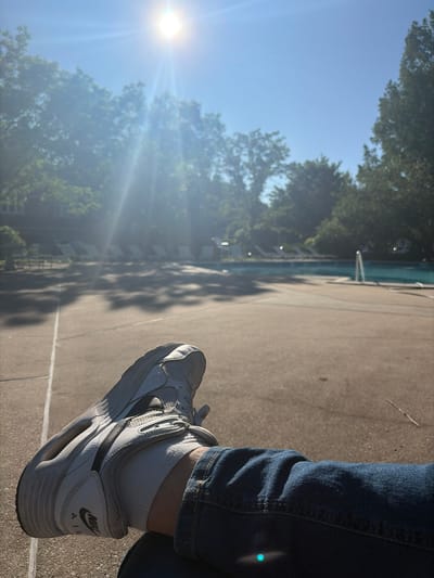 The image depicts a person sitting outdoors in Edison, New Jersey, United States. The person is wearing blue jeans, white socks, and white Nike Air shoes. The location appears to be near a pool, which is partially visible in the background. The sun is shining brightly, creating lens flare. Trees surround the pool area. The ground appears to be concrete or paved.