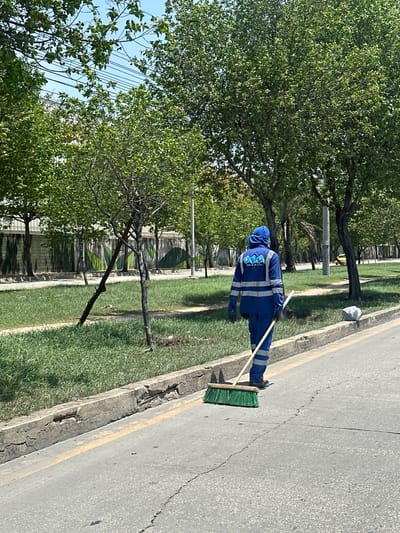 An outdoor, eye-level, medium shot captures a street cleaner in Barranquilla, Colombia, working along the side of a road. The worker, facing away from the camera, is dressed in a bright blue uniform with reflective stripes, including a hooded jacket and pants. They are holding a green-bristled broom with a wooden handle, actively sweeping the asphalt. The road has a yellow painted line along its edge, which is bordered by a broken concrete curb. Beyond the curb is a strip of green grass, dotted with a few sparse trees and some smaller plants. In the background, a row of mature trees with dense green foliage lines the street. A grey concrete wall with a mural-like design featuring green organic shapes is visible behind the trees. The sky is clear and blue, suggesting a sunny day. Further in the background, a yellow car is parked on the right side of the road, and a person on a motorcycle is visible in the distance. The overall environment suggests an urban setting, likely a publicly maintained area within the city.