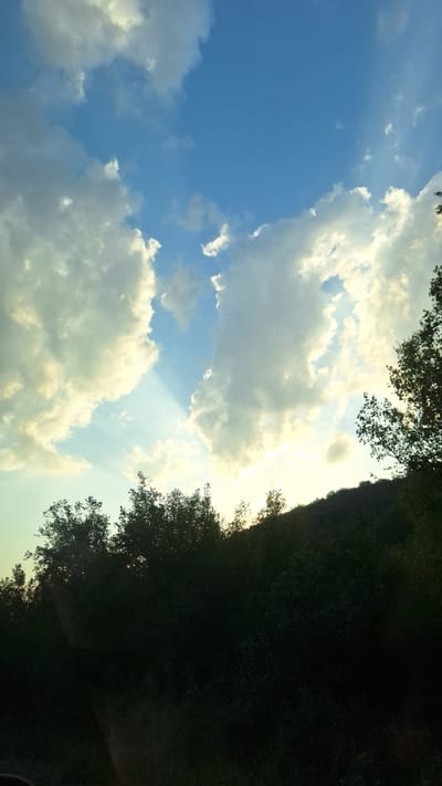 The image shows a view of the sky with clouds and a treeline in Zlatna Panega, Bulgaria. The sky is a mix of blue and white, with sunbeams filtering through the clouds. The sun appears to be low on the horizon, casting a warm light. The treeline is dark and silhouetted against the sky, suggesting it is either early morning or late evening. The ground is not visible in the image, as the camera is pointed upwards. There are no people or animals visible. The overall impression is one of natural beauty and tranquility.