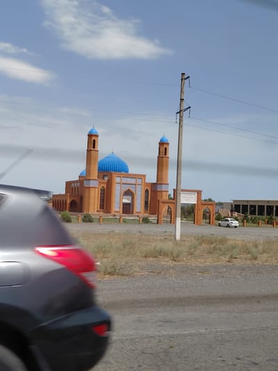 A mosque with a large blue dome and two smaller blue domes, flanked by minarets, is visible in the background under a partly cloudy sky. The mosque's facade is constructed of orange brick with blue tile accents. An archway entrance with a sign reading "ОЛ АНА" is positioned in front of the mosque. A white car is parked near the entrance. In the foreground, the rear of a dark-colored vehicle is blurred, suggesting it is in motion. The environment appears to be arid, with dry grass and gravelly terrain. The location is identified as Бұлақты, Kazakhstan.